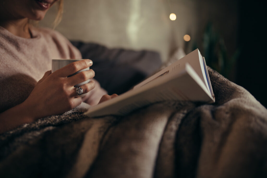 Cropped shot of female hands with book and coffee. Woman reading book on bed at night.